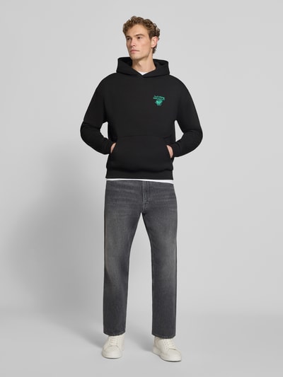 Männermodel trägt Relaxed Fit Hoodie mit Motiv-Print von REVIEW x ANSON´S, Baumwoll-Mix, gerippte Abschlüsse, schwarze Farbe, Ganzkörperansicht von vorne.