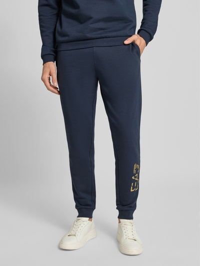 Mężczyzna nosi spodnie dresowe z elastycznym pasem i nadrukiem z logo od EA7 Emporio Armani, bawełna, krój regular fit, długość wewnętrzna 78 cm, widok z przodu na dolną połowę ciała.