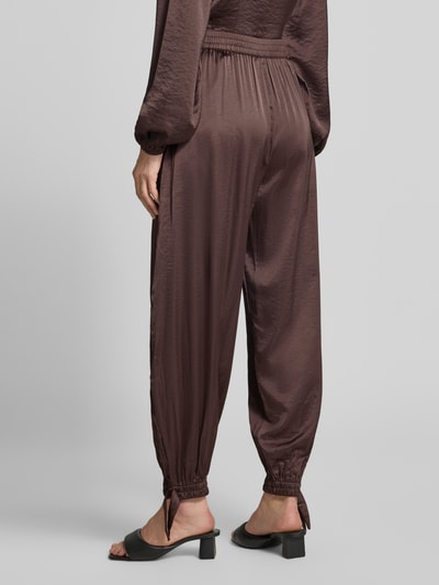 Frauenmodel trägt Loose Fit Stoffhose aus Satin mit Viskose-Anteil von mbyM in Braun, gesmokter Bund, gesmokte Beinabschlüsse mit Schleife, Material-Mix, Ganzkörperansicht Rückansicht.