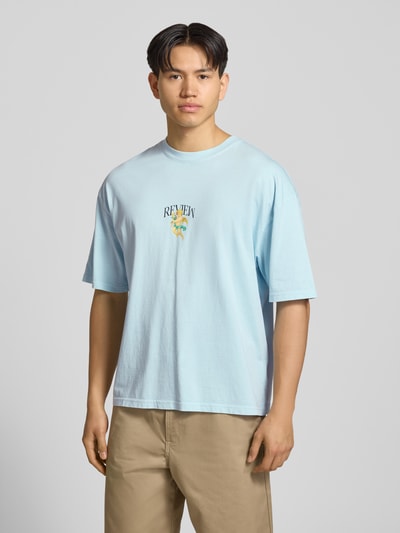 Männermodel trägt T-Shirt mit überschnittenen Schultern von REVIEW in Hellblau, Reine Baumwolle, Lockerer-Schnitt, Label-Print, Ganzkörperansicht von vorne.
