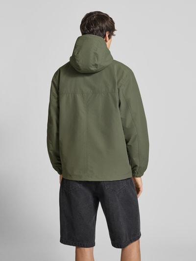 Männermodel trägt Windbreaker mit Label-Stitching von Carhartt Work In Progress in Olivgrün, glatte Struktur, Ganzkörperansicht Rückansicht, Regular Fit, Kapuze, Reißverschluss, Maße Größe M