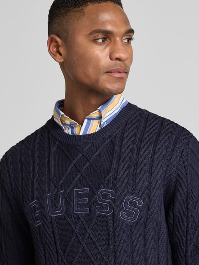 Männermodel trägt dunkelblauen Strickpullover aus Baumwoll-Modal-Mix von Guess mit Rundhalsausschnitt, Nahaufnahme Oberkörper.