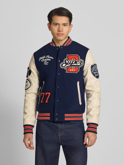 Męskimodel nosi College-Jacke mit Patches und Eingrifftaschen von Superdry, mieszanka bawełny, krój regular fit, stójka, listwa guzikowa, wpuszczane kieszenie, długość tyłu w rozmiarze M: 65 cm, szerokość ramion w rozmiarze M: 45 cm, szerokość klatki piersiowej w rozmiarze M: 49 cm, długość rękawów w rozmiarze M: 70 cm, granatowe i kremowe kolory, kontrastowe naszywki, gładka tekstura, widoczne detale, Ganzkörperansicht von vorne.