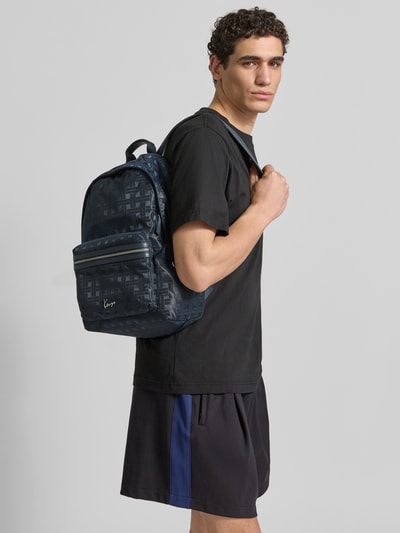 Männermodel trägt Rucksack mit Logo-Applikation von Kenzo in Dunkelblau mit Reißverschluss, Trageriemen, Reißverschlussfach vorne, Steckfach innen, Maße ca. 34 cm Breite, 44 cm Länge, 12 cm Tiefe, Ganzkörperansicht von vorne.