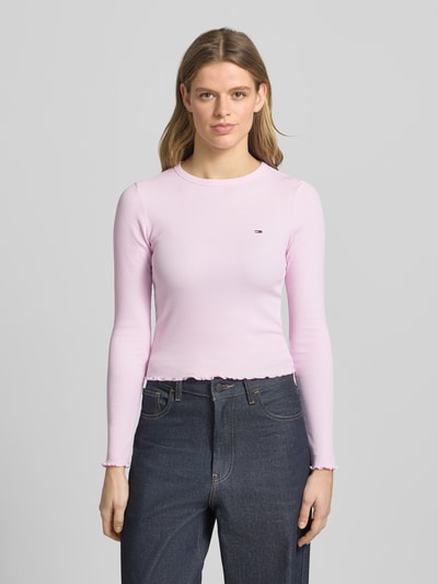 Kobiece modelka nosi Slim Fit Langarmshirt aus Baumwoll-Mix von Tommy Jeans w jasnoróżowym kolorze, gładka faktura, detal z logo, krój slim fit, okrągły dekolt, długi rękaw, drobne prążkowanie materiału, zakończenia rękawów i dołu wykończone falbanką, mieszanka bawełny, widok całej sylwetki z przodu.