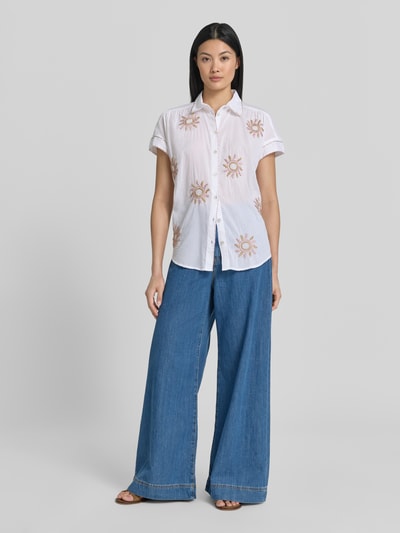 Frauenmodel trägt Regular Fit Bluse im Allover-Look mit Stitchings von Christian Berg Woman aus Reine Baumwolle, Hemdblusenkragen, Knopfleiste, kurze Ärmel, weiße Grundfarbe mit beige Stickereien, dezente Struktur, kombiniert mit blauer Hose, Ganzkörperansicht von vorne.