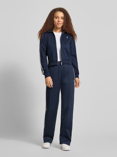 Vrouwenmodel draagt Regular Fit Trackjacke mit Logo-Stitching van Review in donkerblauw met opstaande kraag, met kant, ritssluiting, steekzakken, elastische boorden, ganzkörperaanzicht van voren.