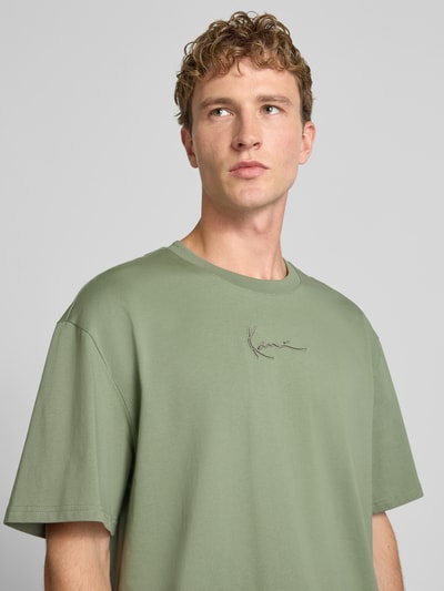 Männermodel trägt T-Shirt mit Logo und Rundhalsausschnitt von Karl Kani in Salbeigrün, reine Baumwolle, Relaxed Fit, Nahaufnahme Oberkörper.