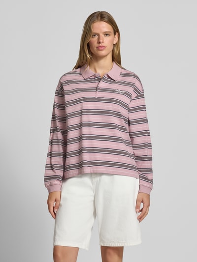 Vrouwenmodel draagt oversized poloshirt met labelstitching model 'Bronson' van Carhartt Work In Progress, puur katoen, roze met witte en grijze strepen, polokraag, ruglengte bij maat S 60 cm, voorkant volledig zichtbaar, Ganzkörperansicht von vorne.