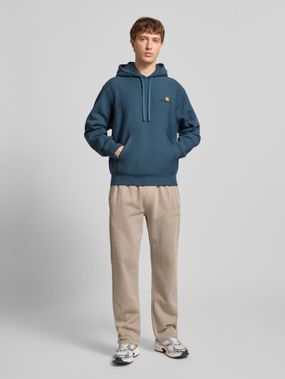 Męskim model nosi Hoodie mit Kapuze od Carhartt Work In Progress, mieszanka bawełny, luźny krój, kaptur, tunel, wyhaftowane logo, kieszeń kangurka, prążkowane wykończenia, długość tyłu dla rozmiaru M: 70 cm, długość rękawa: 64 cm, cała sylwetka z przodu.