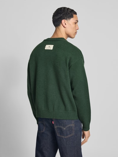 Männermodel trägt Cardigan mit Logo-Stitchings und V-Ausschnitt von ANOTHER COTTON LAB in dunklem Grün, Gerader-Schnitt, Gerippte-Abschlüsse, Rückenlänge 61 cm, Ärmellänge 59 cm, Ganzkörperansicht Rückansicht.