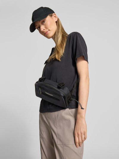 Kobiece modelka nosi Fahrradtasche mit Label-Detail od PEAK PERFORMANCE, kolor czarny, detal z logo, regulowany pasek, główna przegroda na zamek błyskawiczny, boczna przegródka z zamkiem błyskawicznym, szerokość w rozmiarze 1: 26 cm, długość w rozmiarze 1: 14 cm, głębokość w rozmiarze 1: 5 cm, długość paska: 94 cm, zdjęcie całej sylwetki z przodu.