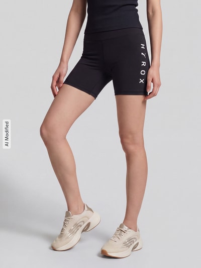 Frauenmodel trägt schwarze Elastische Sportshorts mit Logo-Print Modell 'PUMA X HYROX ESS SHORT' von PUMA, Damen Sportshorts von PUMA Performance, Ganzkörperansicht von vorne, KI-modifiziertes Bild.
