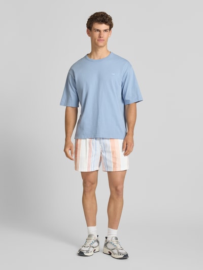 Mannelijk model draagt Relaxed Fit Shorts uit puur katoen van Tommy Jeans met all-over motief, relaxed fit pasvorm, labelpatch, elastische boord met tunnelkoord, zijsteekzakken en dobby materiaalverwerking, kleurige strepen, ganzekeeropname van voren.