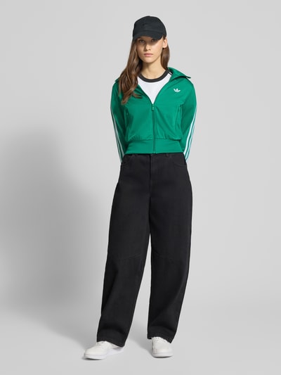 Modelka nosi zieloną Trainingsjacke mit Label-Stitching von adidas Originals, krój regular fit, stójka, zamki, boczne kieszenie, widok całej sylwetki z przodu.