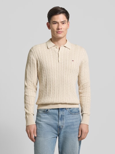 Männermodel trägt Regular Fit Strickpullover aus reiner Baumwolle von Tommy Hilfiger, Optik: Unifarben, Zopfmuster, Logo-Applikation, Polokragen, Langarm, Ärmelabschluss gerippt, Rumpfabschluss gerippt, Materialverarbeitung: Strick, Farbe Beige, Ganzkörperansicht von vorne.