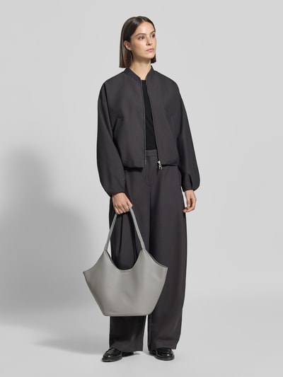 Kobieta modelka trzyma torebkę w stylu Materiał skóropodobny Model 'Nelma' od Seidenfelt, detal z logo, Główna przegroda z zapięciem na magnes, smukły pasek na ramię, Szerokość w rozmiarze 1: 46 cm, Długość w rozmiarze 1: 26 cm, Głębokość w rozmiarze 1: 14 cm, długość paska na ramię w rozmiarze 1: 54 cm, szarość, wyraźna faktura, widok całej sylwetki z przodu