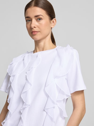Modelka kobieca ma na sobie T-Shirt mit Rüschen und Rundhalsausschnitt od Christian Berg Woman, biała bawełna, prosty krój, z przodu falbany, zbliżenie na górną część ciała.