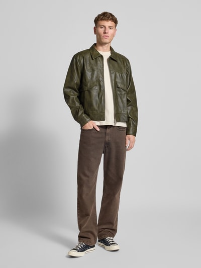 Mężczyzna nosi Jacke in Leder-Optik Modell 'Nerith' von Don't Waste Culture, oliwkowy kolor, prosty krój, kieszenie, cała sylwetka z przodu.