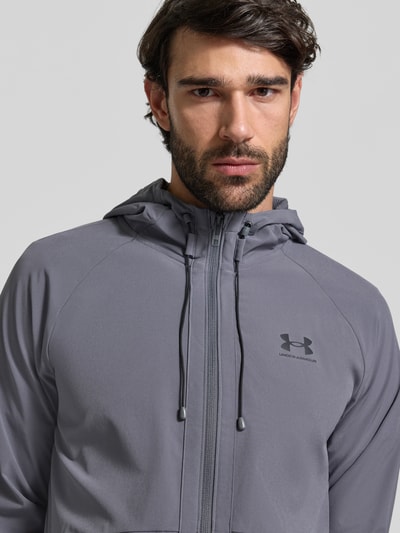 Männermodel trägt Regular Fit Jacke mit Logo-Print Modell 'Vibe' van Under Armour in grijs, capuchon en rits, nahaufnahme oberkörper.