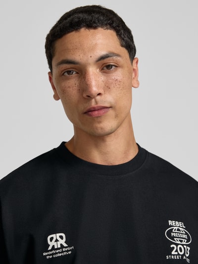 Männermodel trägt schwarzes Herren T-Shirt mit Statement-Print und Rundhalsausschnitt von Redefined Rebel aus Baumwoll-Mix, Nahaufnahme Oberkörper.