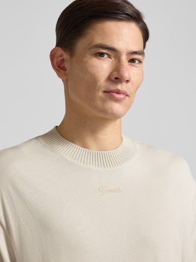 Männermodel trägt T-Shirt mit Label-Detail von Guess in Beige, Reine Baumwolle, Lockerer-Schnitt, Gerippter Rundhalsausschnitt, Nahaufnahme Oberkörper. Rückenlänge bei Größe M: 73 cm.