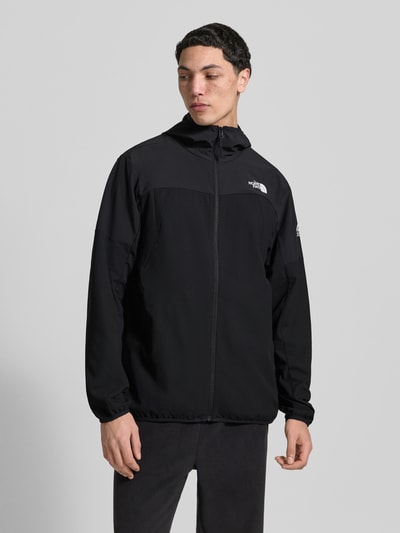 Męski model nosi Jacke mit Logo und Reißverschluss od The North Face, czarna mieszanka materiałów z dodatkiem streczu, krój regular fit, logo na piersi, wysokie zapięcie z kapturem, zamek błyskawiczny, długość tyłu w rozmiarze M: 63 cm, szerokość ramion w rozmiarze M: 48 cm, szerokość klatki piersiowej w rozmiarze M: 33 cm, długość rękawów w rozmiarze M: 65 cm, widok całej sylwetki z przodu.