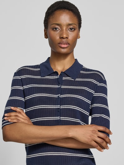 Frauenmodel trägt Slim Fit Poloshirt aus reiner Wolle von Closed in Dunkelblau mit weißen Streifen, Halbarm, Polokragen, Nahaufnahme Oberkörper.
