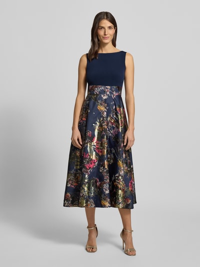 Frauenmodel trägt Abendkleid mit U-Boot-Ausschnitt von Christian Berg Cocktail, Material-Mix mit Stretch-Anteil, taillierter Schnitt, floraler Rock, blickdichter Unterrock, Nahtreißverschluss, Ganzkörperansicht von vorne.