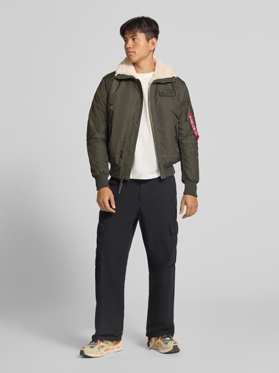Männermodel trägt Bomberjacke mit Label-Patch und Kentkragen von Alpha Industries in Olivgrün, Teddyfell-Kragen, Reißverschluss, Ganzkörperansicht von vorne. Rückenlänge 65 cm, Schulterbreite 45 cm, Brustweite 56 cm, Ärmellänge 65 cm, Regular Fit.