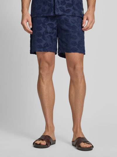 Męski model nosi Relaxed Fit Shorts aus Frottee mit Gesäßtasche od Scotch & Soda, mieszanka bawełny, krój relaxed fit, frota, elastyczny pas, wpuszczane kieszenie, kieszeń z tyłu, detal z logo, szerokość w rozmiarze M: 41 cm, długość wewnętrzna nogawki w rozmiarze M: 18 cm, ciemnoniebieski, faktura froty, liściasty wzór, widok całej sylwetki z przodu.