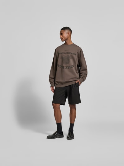 Männermodel trägt Oversized Sweatshirt mit Logo-Print von MM6 Maison Margiela in Braun, reine Baumwolle, elastische Abschlüsse, Ganzkörperansicht von vorne.
