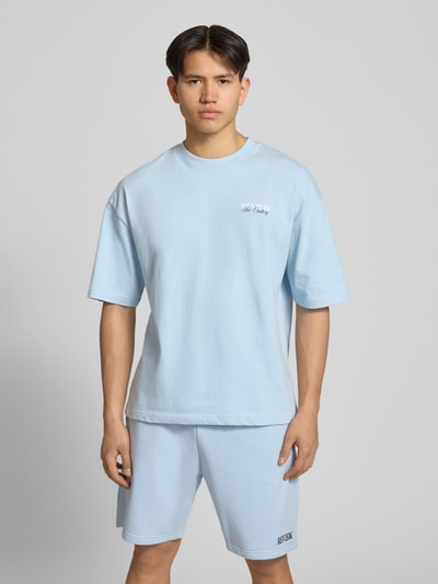 Männermodel trägt T-Shirt mit Label-Detail von REVIEW in Hellblau, lockerer Schnitt, reine Baumwolle, Halbarm, Ganzkörperansicht von vorne.