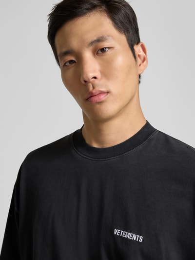 Männermodel trägt Relaxed Fit T-Shirt aus reiner Baumwolle von VETEMENTS in Schwarz mit Rundhalsausschnitt und Label-Stitching, Nahaufnahme Oberkörper.