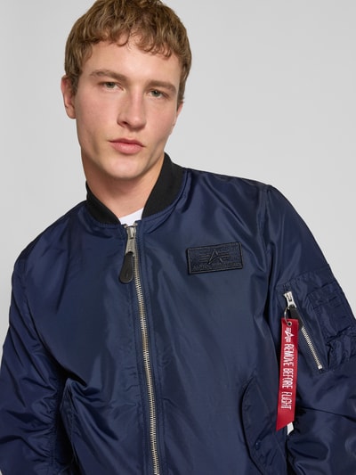 Męski model nosi Regular Fit Bomberjacke mit Logo-Patch od Alpha Industries, granatowy kolor, gładka tekstura, krój regular fit, stójka, kieszenie boczne, zamek błyskawiczny, kieszeń na rękawie z zamkiem, długość tyłu w rozmiarze M: 65 cm, szerokość ramion w rozmiarze M: 45 cm, szerokość klatki piersiowej w rozmiarze M: 54 cm, długość rękawa w rozmiarze M: 69 cm, zbliżenie na górną część tułowia