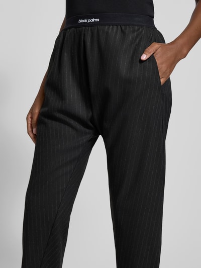 Frauenmodel trägt Stoffhose mit elastischem Logo-Bund von black palms, schwarz mit feinen Nadelstreifen, lockerer Schnitt, Nahaufnahme Oberkörper.