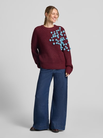 Frauenmodel trägt Relaxed Fit Pullover mit Woll-Anteil Modell 'KOSTAR' von Essentiel Antwerp in Dunkelrot mit blauen Applikationen, weite Jeans, Ganzkörperansicht von vorne. Damen Pullover von Essentiel Antwerp.