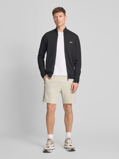 Männermodel trägt schwarze Jacke, weißes T-Shirt, beige Shorts und Sneakers, gedeckte Farben, glatte Texturen, Ganzkörperansicht von vorne.
