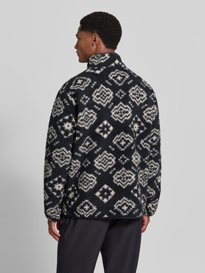 Męski model ma na sobie sweter Regular Fit Pullover mit Logo-Patch od Columbia, Polar, krój regular fit, naszywka z logo, kołnierz zapinany, Długość tyłu w rozmiarze M: 70 cm, Długość rękawów w rozmiarze M: 68 cm, czarne i kremowe geometryczne wzory, widok całej sylwetki z tyłu.