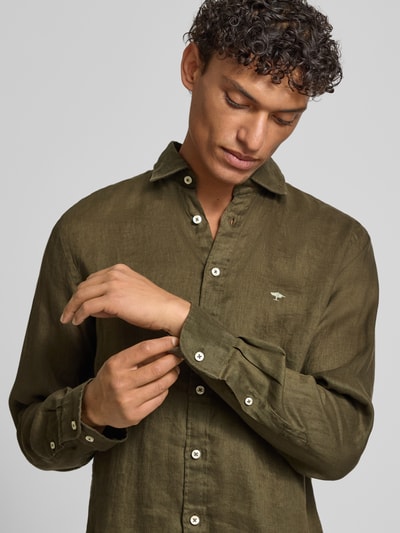 Męski model nosi Slim Fit Leinenhemd mit Haifischkragen od Fynch-Hatton z lnu, krój slim fit, wyhaftowane logo, kołnierzyk typu cutaway, długość tyłu w rozmiarze M 77 cm, szerokość ramion w rozmiarze M 47 cm, długość rękawów w rozmiarze M 61 cm, jednolity oliwkowy kolor, gładka faktura, zbliżenie na górną część ciała.