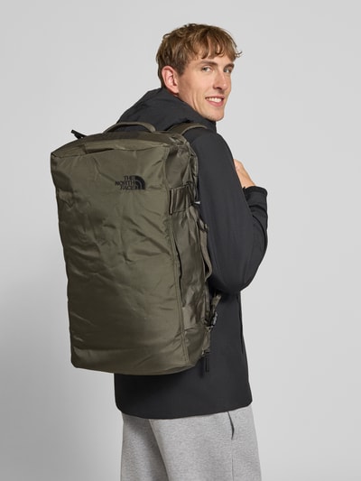 Männermodel trägt Duffel Bag mit Logo-Print Modell 'Voyager' von The North Face in Olivgrün, glatte Textur, Ganzkörperansicht Rückansicht, Herren Duffel Bag, Hauptfach mit Reißverschluss, verstellbare Schulterriemen, Innenfächer mit Reißverschluss, 30x20x9 cm, Schulterriemen 13 cm.