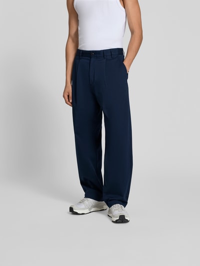 Männermodel trägt Wide Fit Stoffhose mit Bundfalten von Forét in Dunkelblau, elastischer Baumwoll-Mix, Ganzkörperansicht von vorne.