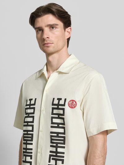 Männermodel trägt Regular Fit Freizeithemd mit Label-Print Modell 'Banks' von WOODBIRD, cremefarben, Kentkragen, Nahaufnahme Oberkörper. Rückenlänge 76 cm, Schulterbreite 52 cm, Ärmellänge 28 cm bei Größe M.