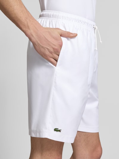 Mężczyzna nosi Relaxed Fit Sport-Shorts mit Logo-Stitching von Lacoste Sport, białe, jednokolorowe, elastyczny pas z troczkiem, boczne kieszenie, aplikacja logo, wykonane z gładkiego materiału, zbliżenie na prawy bok i kieszeń, detal.