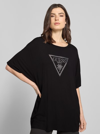 Modelka nosi T-Shirt mit Logo-Print Modell 'COULISSE' od Guess z elastycznej wiskozy, czarne, luźny krój, widoczny nadruk, zdjęcie: ujęcie całej sylwetki z przodu.