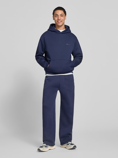 Männermodel trägt Hoodie mit Logo-Print von REVIEW in Dunkelblau, lockere Passform, Kängurutasche, Baumwoll-Mix, Ganzkörperansicht von vorne