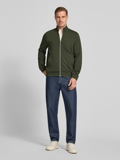 Männermodel trägt Sweatjacke mit Stehkragen und Eingrifftaschen von PAUL SMITH aus Baumwoll-Mix mit Modal-Anteil in Dunkelgrün, Regular-Fit, Reißverschluss, kombiniert mit Jeans und hellen Sneakern, Ganzkörperansicht von vorne.