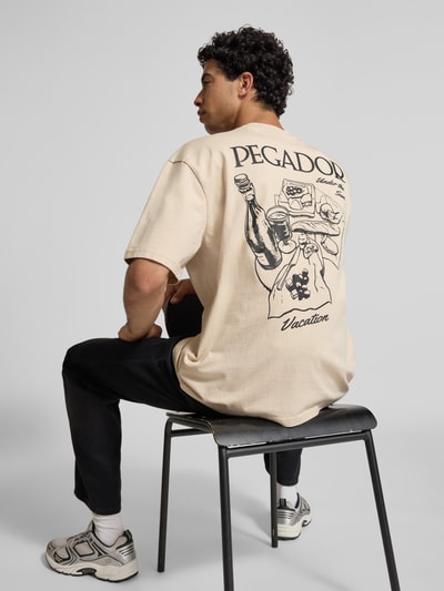 Männermodel trägt Oversized T-Shirt mit Label-Print Modell 'Paide' von Pegador, beige, katoen, rugprint, heren, rugaanzicht, context: heren T-shirt van Pegador, pure katoen, oversized fit, model 'Paide', ronde hals, labelprint, ruglengte maat M: 70 cm.