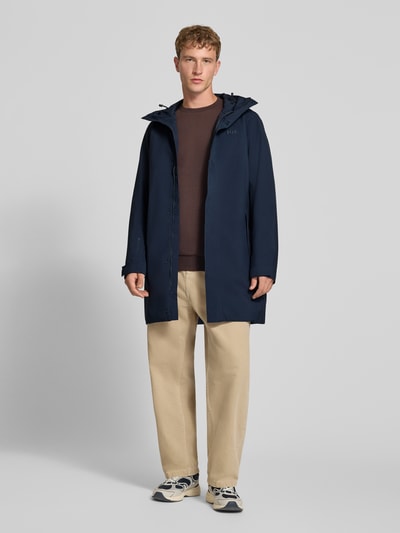 Männermodel trägt Regular Fit Mantel mit Kapuze Modell 'Brussel' von Helly Hansen in Dunkelblau, glatte Textur, Ganzkörperansicht von vorne. Rückenlänge bei Größe M: 96 cm, Schulterbreite: 46 cm, Brustweite: 51 cm, Ärmellänge: 66 cm, Logo-Detail.