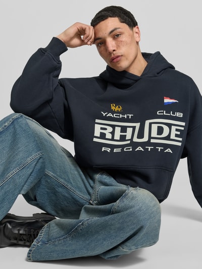 Männermodel trägt Regular Fit Hoodie mit Logo-Print von RHUDE aus reiner Baumwolle, Känguru-Tasche, elastische Abschlüsse, Ganzkörperansicht von vorne. Rückenlänge bei Größe M: 65 cm.